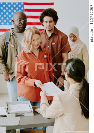 Vertical medium long shot of ethnically diverse men and women standing in line at voting place taking ballot papers 115735367