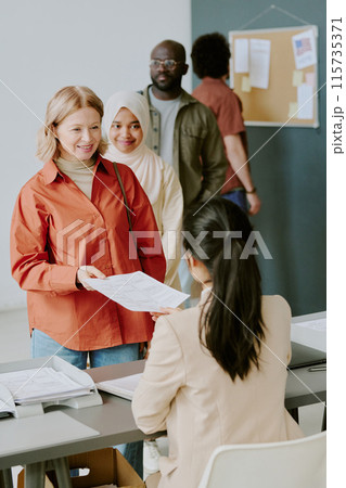 Vertical shot of mature Caucasian woman taking ballot paper at polling station to vote for her favorite candidate Vertical shot of mature Caucasian woman taking ballot paper at polling station to vote for her favorite candidate 115735371
