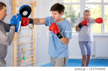 Guy in boxing gloves practicing punches on mitts with coach during self-defense course 115743600