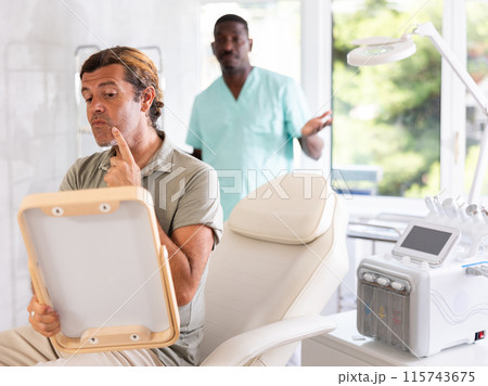 Man dissatisfied with results of rejuvenating at cosmetologist and meticulously examines reflection Man dissatisfied with results of rejuvenating at cosmetologist and meticulously examines reflection 115743675