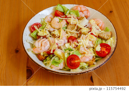 Fresh healthy caesar salad with prawns, cherry tomatoes, lettuce, parmesan cheese, dressing and croutons on wooden table 115754492