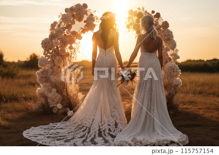 Couple of brides wearing wedding dresses are holding hands in the background of romantic wedding arch at the sunset field. Same sex wedding. Couple of brides wearing wedding dresses are holding hands in the background of romantic wedding arch at the sunset field. Same sex wedding. 115757154