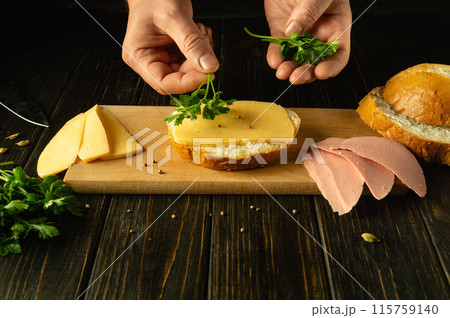 Making sandwiches with cheese and parsley. Close-up hands of a chef on the kitchen table preparing breakfast. Place for advertising on a dark background 115759140
