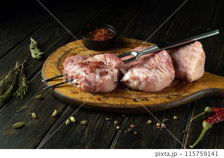 Preparing juicy beef steaks for the grill. Sliced raw meat pieces and a fork on a cutting board. Meat spices are scattered on the kitchen table Preparing juicy beef steaks for the grill. Sliced raw meat pieces and a fork on a cutting board. Meat spices are scattered on the kitchen table 115759141