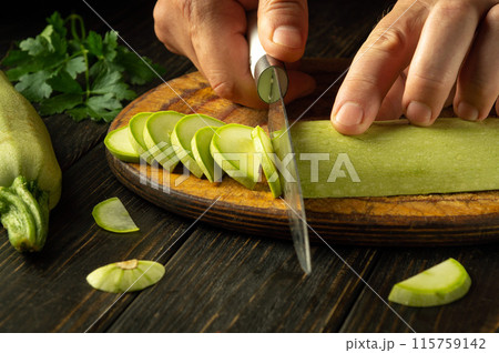 Slicing zucchini with a knife and hands of a chef on a cutting board to prepare a vegetable salad on the kitchen table Slicing zucchini with a knife and hands of a chef on a cutting board to prepare a vegetable salad on the kitchen table 115759142