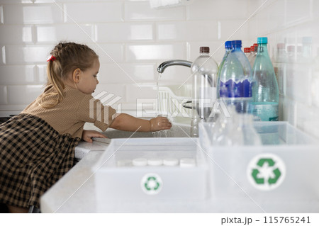 The little girl washes a plastic bottle in the sink and then puts it in a box with a recycling sign. A child helps clean plastic bottles before they are sent to the recycling service. The little girl washes a plastic bottle in the sink and then puts it in a box with a recycling sign. A child helps clean plastic bottles before they are sent to the recycling service. 115765241