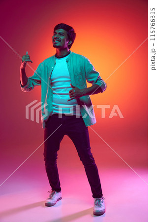 Overjoyed young Arabian man wearing casual clothes and dancing of joy and happiness in neon light against gradient studio background. 115766315