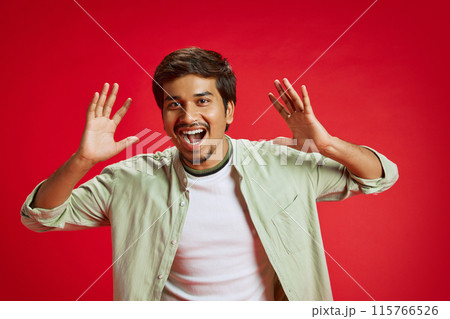 Portrait of emotional young Indian man with excited facial expression looking at camera against red studio background. 115766526