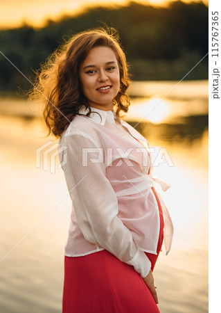 Beautiful young pregnant woman in red dress at sunset on the riverbank 115767365