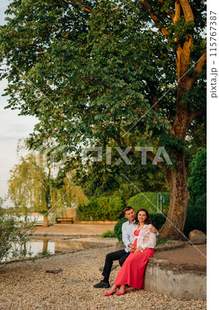 man and pregnant woman in red dress at sunset on the riverbank 115767387