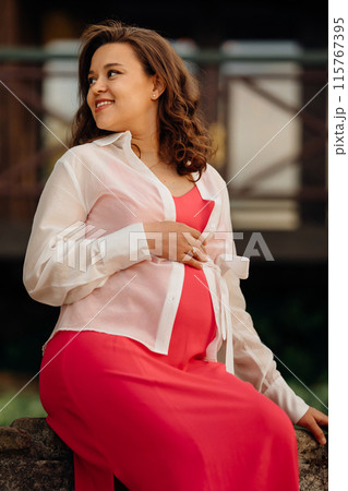 Beautiful young pregnant woman in red dress sitting on in park. 115767395