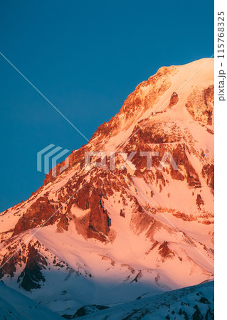Georgia. Mount Kazbek Covered Snow In Winter Sunrise. Morning Dawn Colored Top Of Mountain In Pink-orange Colors. Awesome Winter Georgian Nature Landscape. Georgia. Mount Kazbek Covered Snow In Winter Sunrise. Morning Dawn Colored Top Of Mountain In Pink-orange Colors. Awesome Winter Georgian Nature Landscape. 115768325