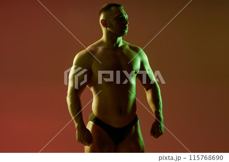 Medium full shot of confident athletic man shirtless showing upper body muscles with hard chest and arm muscles on red background. Studio image of sportsman with contour green and neutral light 115768690