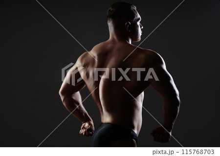 Medium backlight image of sexy brutal male model sideways to camera showing flexed back muscles in dark studio with dramatic light on black background, copy space Medium backlight image of sexy brutal male model sideways to camera showing flexed back muscles in dark studio with dramatic light on black background, copy space 115768703