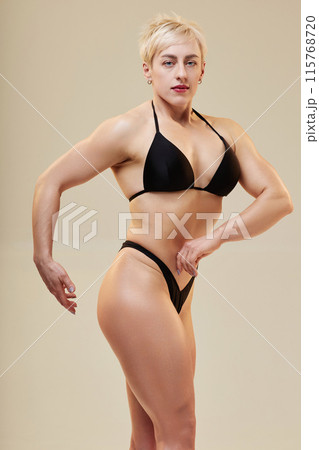 Medium full shot of attractive lady bodybuilder showing off chest and arm muscles isolated in studio on neutral background 115768720