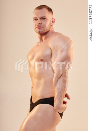 Medium long shot portrait of confident bodybuilder performing side triceps pose topless. Handsome sportsman showing arm and chest muscles looking at camera isolated on neutral background 115768773