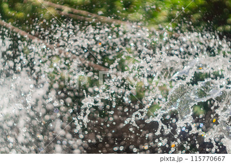 Drops of water flying in the fountain of a city park against the background of a blurred green background of tree foliage. Park areas, summer days with hot weather, urban recreation areas. 115770667