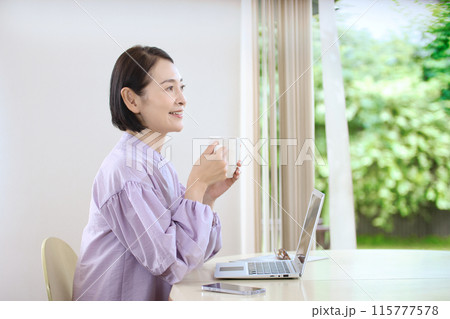 リビングでコーヒーを飲みながらパソコンを見るミドル女性 115777578
