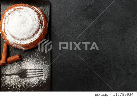 Round, delicious bun with cinnamon and powdered sugar on a black plate on a gray background Round, delicious bun with cinnamon and powdered sugar on a black plate on a gray background 115789934