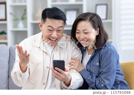 Happy young Asian couple, man and woman are sitting on the sofa at home and are happy with the received news and message they are reading on the phone. 115791098