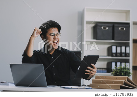 Celebrating success. Young asian business man working with laptop, tablet and papers on desk at office. He feeling good and happy. 115795932