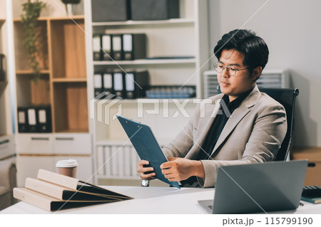 Tired young Asian man, accountant, businessman, freelancer. He sits in the office at a table with a laptop, looks through documents, holds his head in his hands, sighs a hard and boring day. Tired young Asian man, accountant, businessman, freelancer. He sits in the office at a table with a laptop, looks through documents, holds his head in his hands, sighs a hard and boring day. 115799190