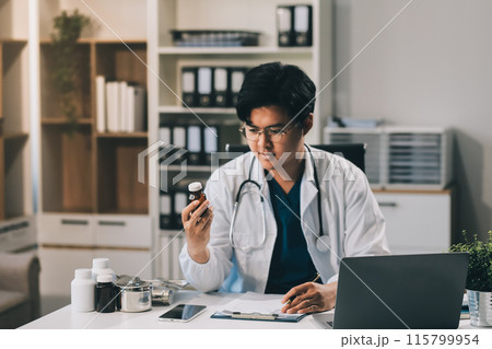 .Handsome young doctor with a beard and a lab coat taking of his glasses to read an email in his laptop .Handsome young doctor with a beard and a lab coat taking of his glasses to read an email in his laptop 115799954