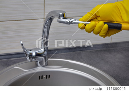 A plumber in yellow rubber gloves unscrews the faucet aerator with an adjustable wrench 115800043