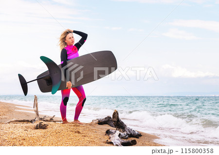young woman walks along the water on the shore of the beach to do the water sport of hydrofoil surfing. Sexy sportswoman holding hydrofoil in her hands young woman walks along the water on the shore of the beach to do the water sport of hydrofoil surfing. Sexy sportswoman holding hydrofoil in her hands 115800358