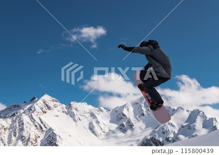 Portrait of a young man snowboarder jumping on a snowboard in sportswear, against the backdrop of high snow-capped mountains and sky 115800439