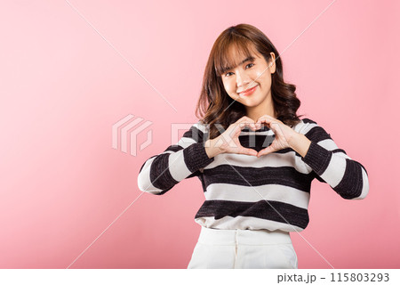 A confident woman smiles and creates a heart symbol with two hands on a pink background. Asian portrait of a beautiful young female expressing love and happiness for Valentine's Day. A confident woman smiles and creates a heart symbol with two hands on a pink background. Asian portrait of a beautiful young female expressing love and happiness for Valentine's Day. 115803293