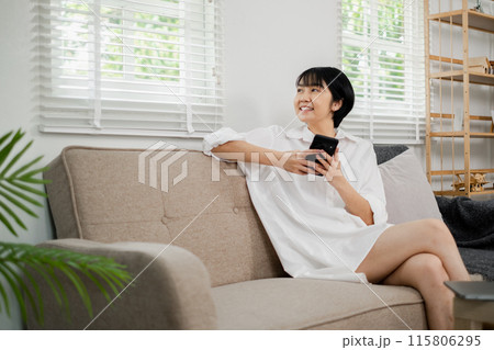 Young woman in a white dress sitting on a sofa, holding a smartphone, smiling, in a bright and modern living room with natural light and minimalist decor. Young woman in a white dress sitting on a sofa, holding a smartphone, smiling, in a bright and modern living room with natural light and minimalist decor. 115806295