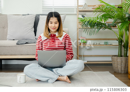 Young woman sitting on the floor using a laptop in a cozy living room with modern decor and indoor plants. Young woman sitting on the floor using a laptop in a cozy living room with modern decor and indoor plants. 115806303