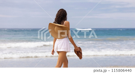 Woman Walking on Beach Holding Sandals and Sun Hat on Shoulder 115806440