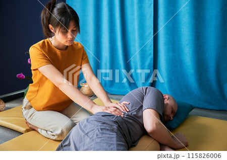 Masseuse making thai yoga massage. Woman therapist doing traditional massage treatment for male patient indoors. Bald client lying on mat, meditating, enjoying hand massage in calm atmosphere. 115826006