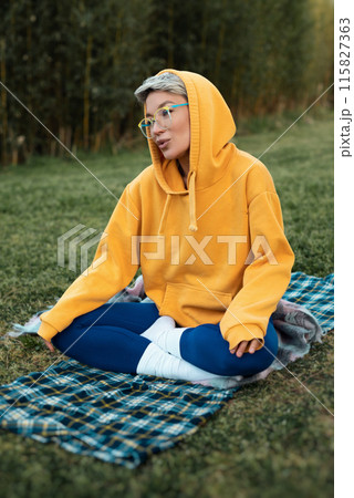 cute girl with glasses in an orange hoodie is sitting in a park in nature expressing kind emotions cute girl with glasses in an orange hoodie is sitting in a park in nature expressing kind emotions 115827363