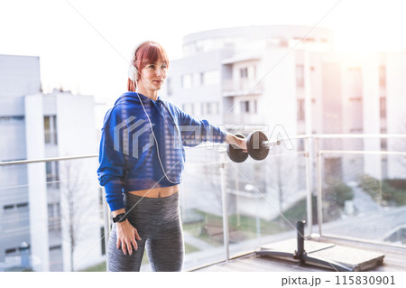 Redhead woman's home workout routine with dumbbell at balcony. Home fitness workout with view on city. Sporty woman wearing warm sportswear. 115830901