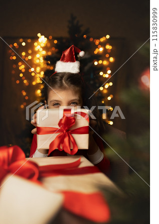 Happy 6 years old girl in Santa Claus costume peeks out from behind gifts boxes with red ribbons and festive lights on background 115830999