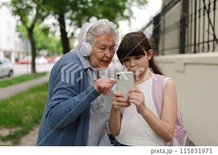 Grandma picking up young girl from school at afternoon. Granddaughter showing funny video on smartphone to senior grandmother. 115831971