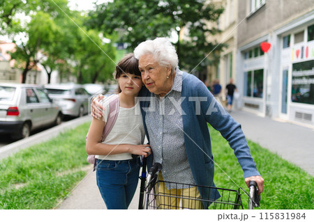 Grandma with walker picking up young girl from school at afternoon. Granddaughter spending time with senior grandmother outdoors in city after school. 115831984