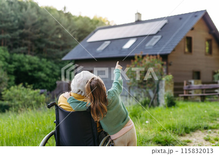 Granddaughter showing grandma solar panels on roof. Senior lady in wheelchair spending time in nature. Granddaughter showing grandma solar panels on roof. Senior lady in wheelchair spending time in nature. 115832013