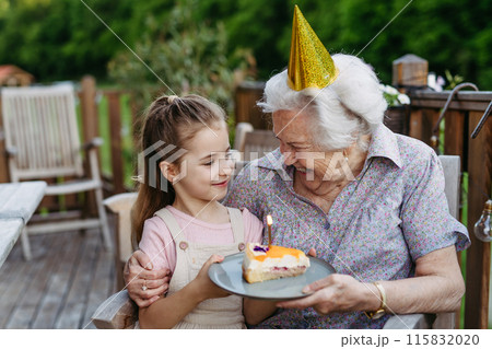 Granddaughter celebrating birthday with elderly grandma, holding cake with candle. Senior lady spending time with young girl, enjoying together time. 115832020