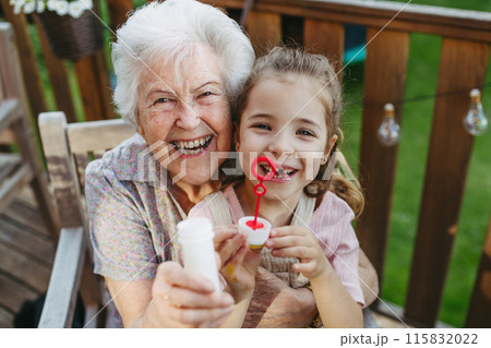 Granddaughter blowing bubbles with elderly grandma. Senior lady spending time with young girl, enjoying together time. 115832022