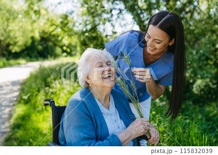 Female caregiver and senior woman in wheelchair picking wild flowers. Nurse and elderly woman enjoying a warm day in nursing home, public park. Female caregiver and senior woman in wheelchair picking wild flowers. Nurse and elderly woman enjoying a warm day in nursing home, public park. 115832039