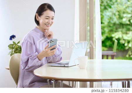 リビングでスマホを操作しながらパソコンを見るミドル女性 115834280