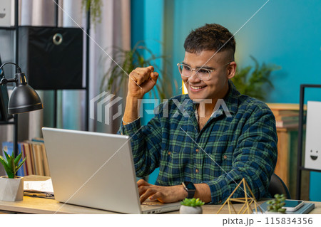 Overjoyed Indian man working on laptop celebrate success win money in lottery get online good news Overjoyed Indian man working on laptop celebrate success win money in lottery get online good news 115834356