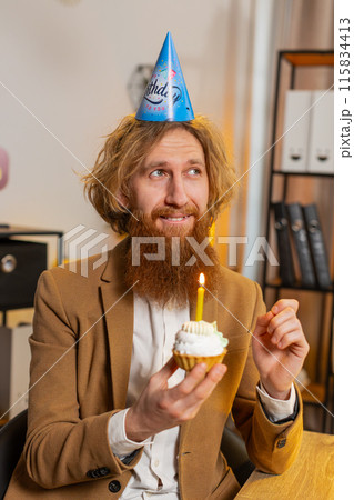 Happy business office man wears festive birthday hat hold cupcake makes wish blowing burning candle 115834413