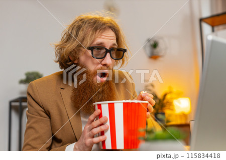 Business man taking break from work wearing 3D glasses earing popcorn watching movie on laptop Business man taking break from work wearing 3D glasses earing popcorn watching movie on laptop 115834418