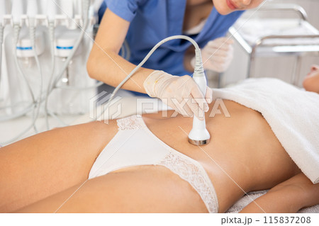 Young female cosmetologist performs ultrasound massage on belly of young woman Young female cosmetologist performs ultrasound massage on belly of young woman 115837208