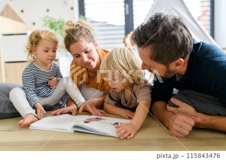 Young nuclear family playing with toys in a living room. Parents and children lying on floor, looking at children's story book, spending weekend day indoors. 115843476
